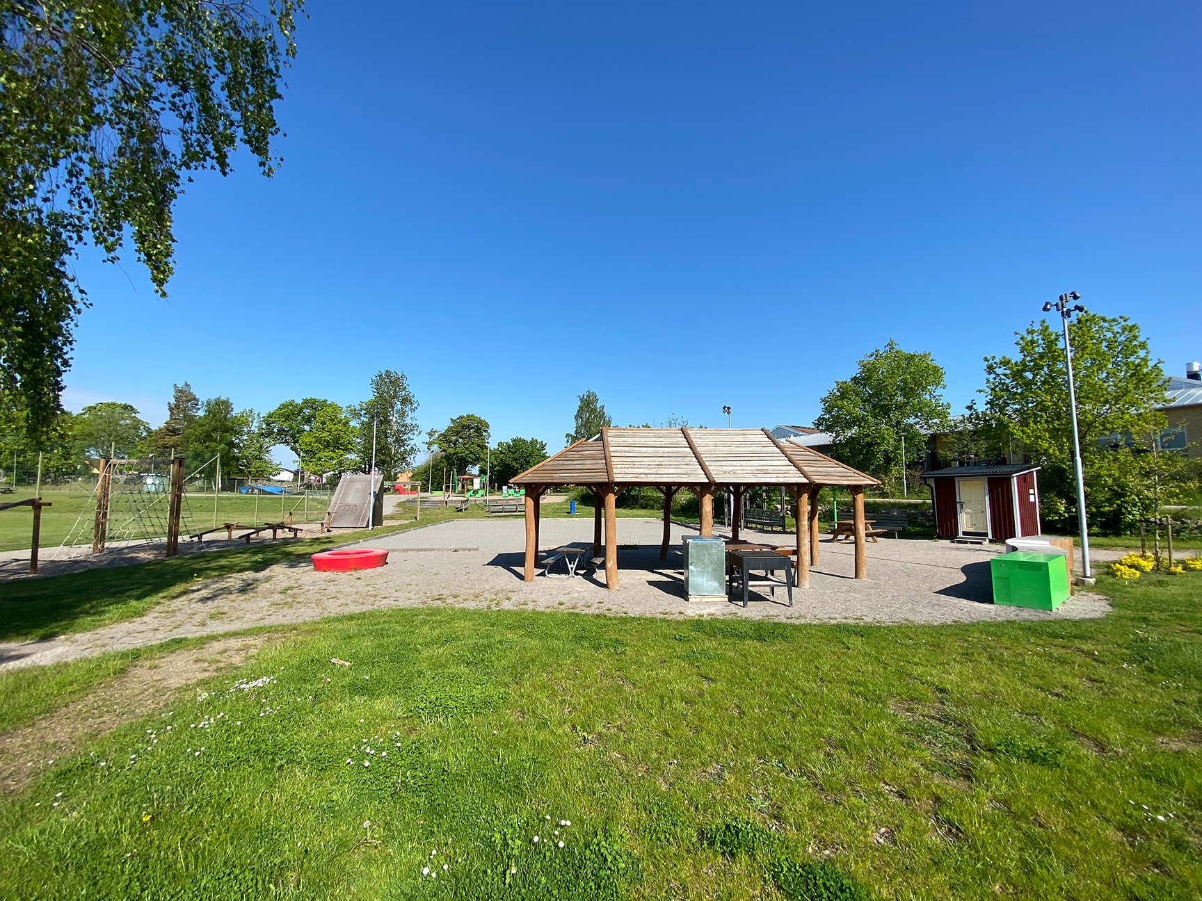 Solrik dag i parken med grillhytte, hinderløype og grønt gress.