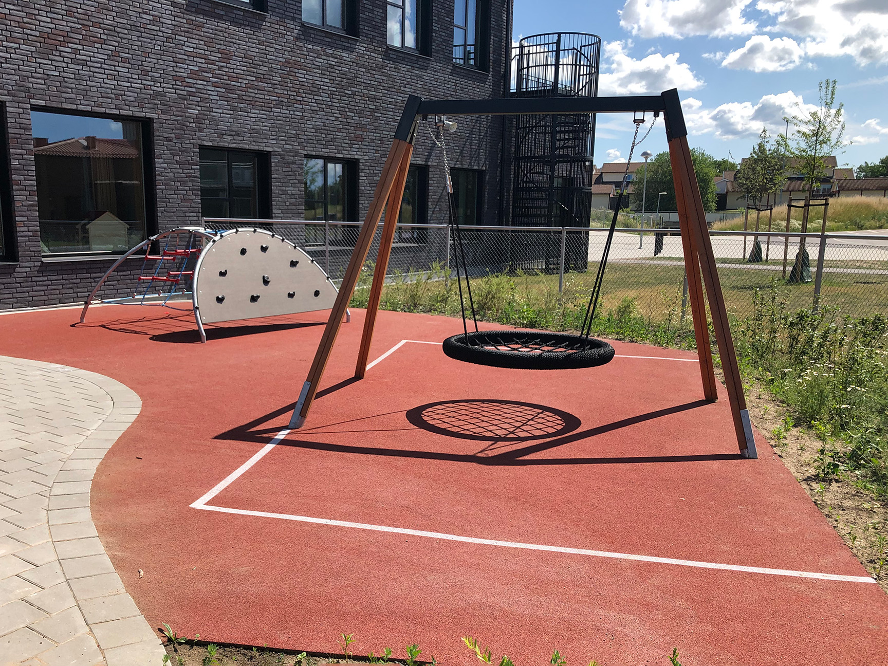 Huske og klatrestativ på rødt gummidekke foran en skolebygning.
