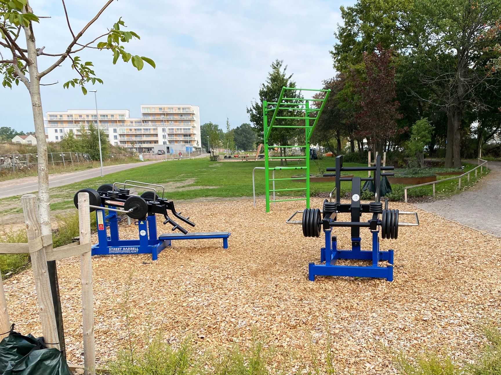 To blå treningsapparater og en grønn treningsstasjon i en park med barkflis som underlag