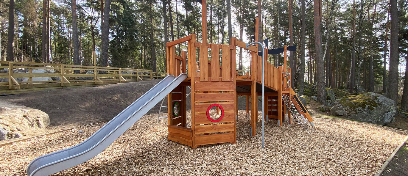 Lekeapparat i tre med rutsjebane og brannmannstang, plassert i et skogsområde.